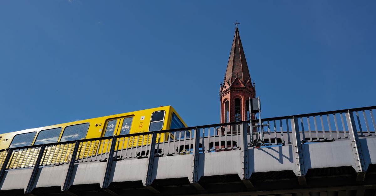 A vibrant yellow train crosses a bridge with a historic church spire in the background against a clear blue sky in Berlin.